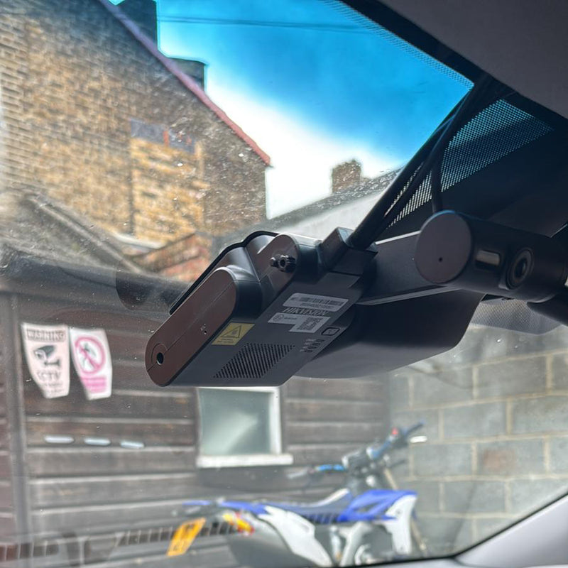 A black car dash cam mounted on a windshield bracket inside a vehicle, with a brick building and a blue motorcycle visible through the window.